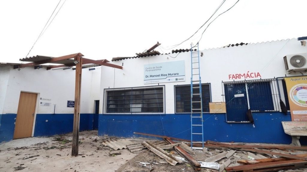 Fachada de prédio com parede azul e branca, placa de Farmácia, janelas e escada ao centro, com materiais e entulho no chão; visão externa em ambiente urbano.