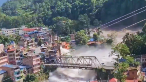 Vista aérea de uma cidade em encosta com ruas e casas, e uma ponte de metal atravessando o rio enquanto uma grande nuvem de poeira e fumaça se espalha pela área