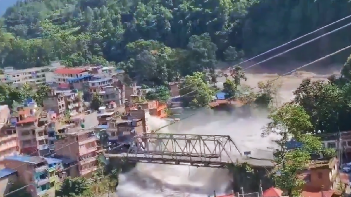 Vista aérea de uma cidade em encosta com ruas e casas, e uma ponte de metal atravessando o rio enquanto uma grande nuvem de poeira e fumaça se espalha pela área