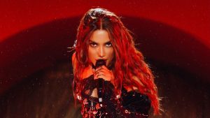 Ensaios da Anitta: Anitta aparece no palco cantando ao microfone, com cabelos longos ruivos e iluminação vermelha, em divulgação de datas da turnê 2027. Foto: Reprodução/Instagram @anitta