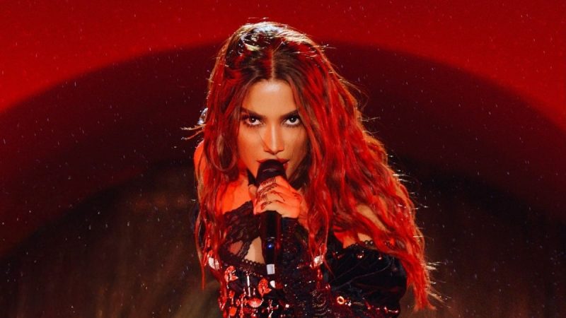 Ensaios da Anitta: Anitta aparece no palco cantando ao microfone, com cabelos longos ruivos e iluminação vermelha, em divulgação de datas da turnê 2027. Foto: Reprodução/Instagram @anitta