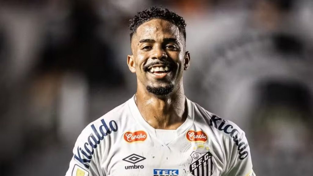 Hayner em foto de futebol com uniforme branco e detalhes de patrocinadores, sorrindo em campo durante partida, com fundo desfocado em estádio.
