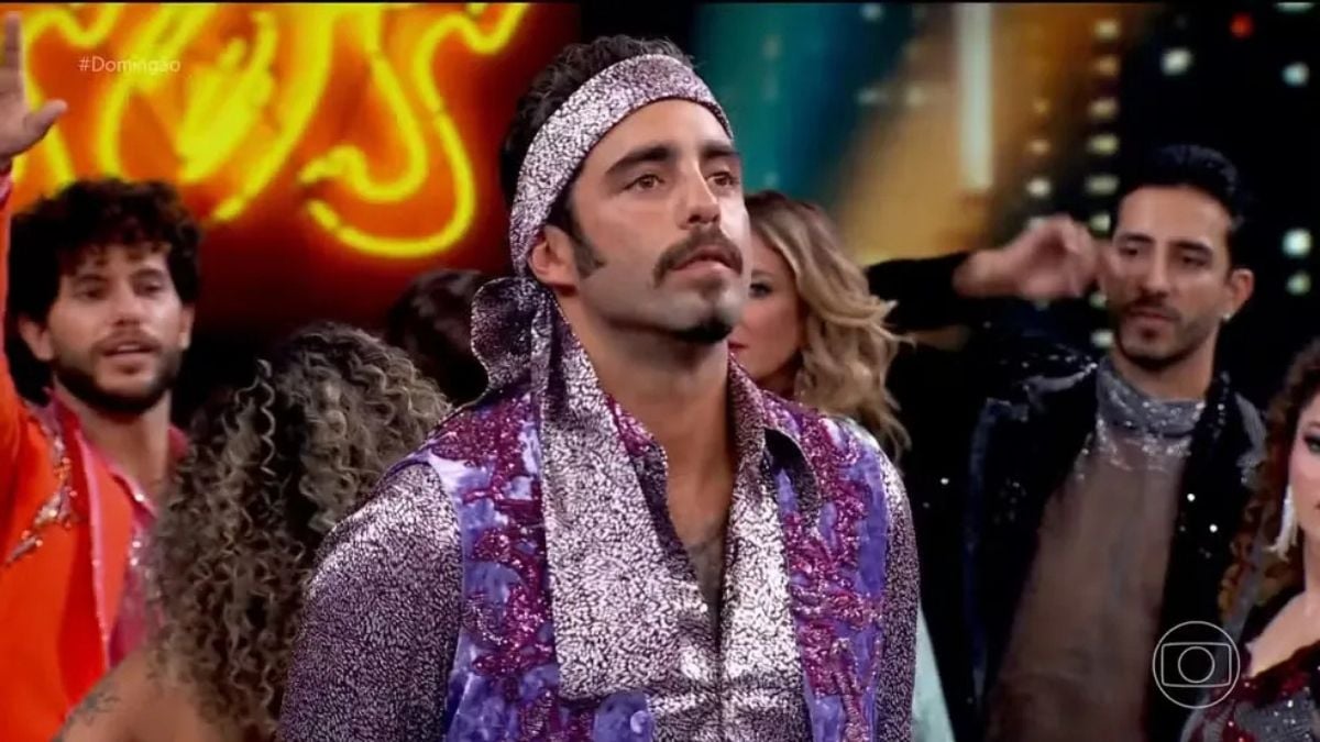 Pedro Scooby aparece “congelado” durante uma apresentação na Dança dos Famosos, em cenário de palco com outros participantes ao fundo, revivendo um meme do BBB 22.