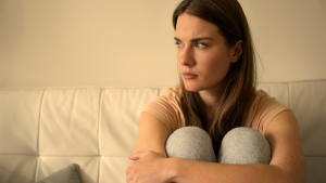 Mulher jovem sentada em sofá, abraçando os joelhos e olhando para o lado em expressão pensativa e melancólica, em ambiente interno com luz suave.