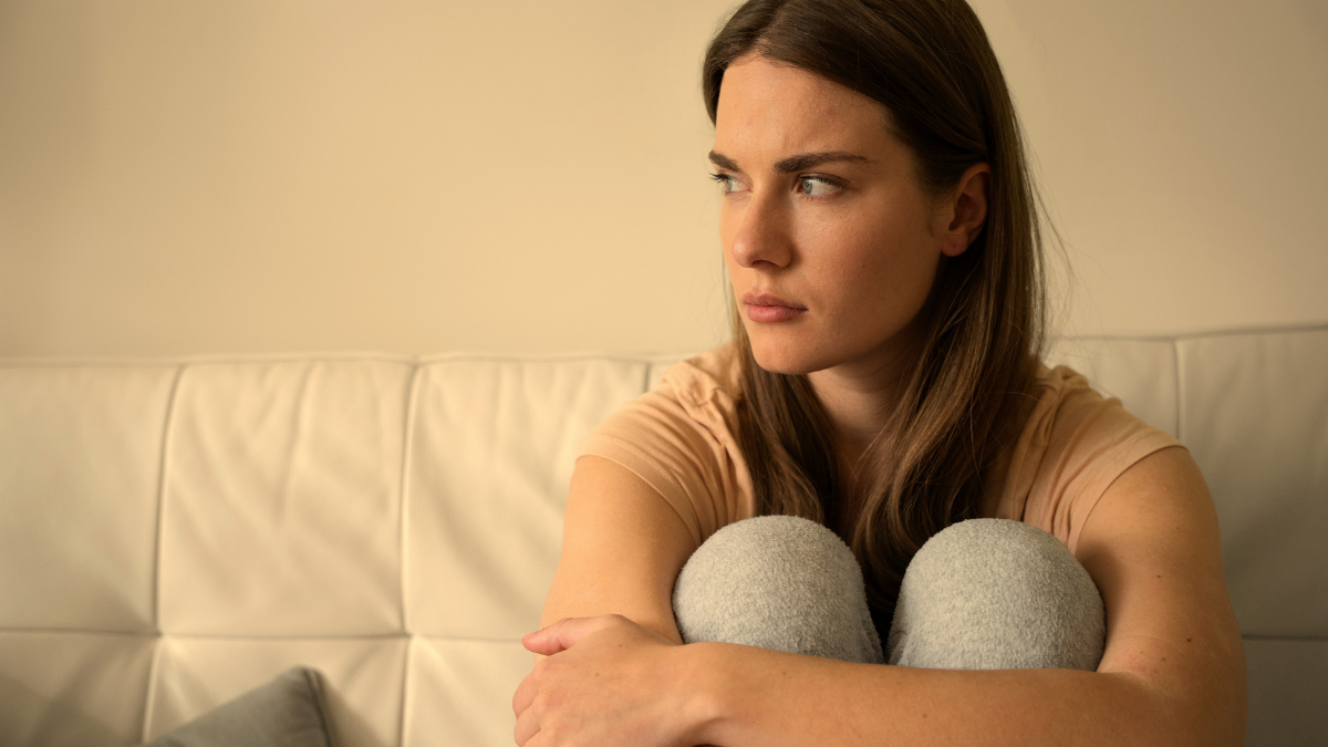 Mulher jovem sentada em sofá, abraçando os joelhos e olhando para o lado em expressão pensativa e melancólica, em ambiente interno com luz suave.