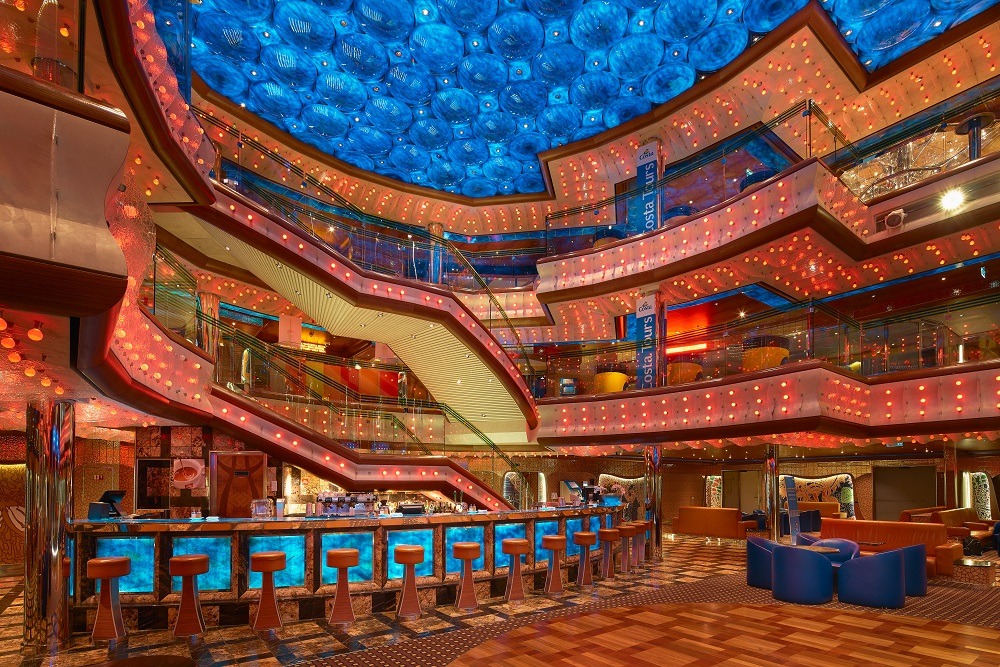 temporada de cruzeiros 2026/2027: Interior de um navio Costa Diadema, com bar ao fundo, balcões iluminados por luzes decorativas, poltronas e um teto em azul com detalhes de iluminação, ambiente de cruzeiro