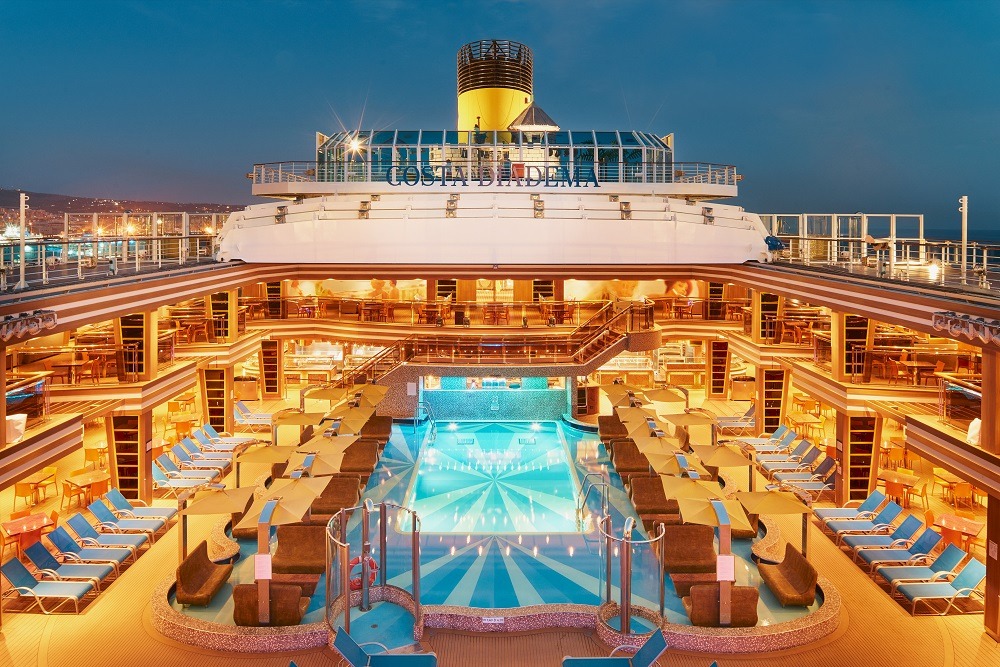 Vista noturna da piscina e do deck do navio Costa Diadema, com espreguiçadeiras e luzes refletindo no mar, em anúncio da temporada de cruzeiros 2026/2027.