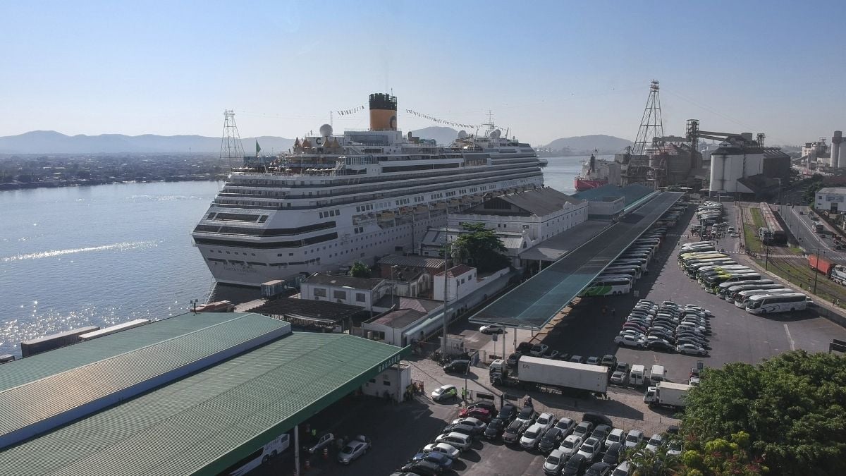 Vista aérea de um navio de cruzeiro atracado no porto de Santos, com carros estacionados e cais ao lado do mar, destacando a temporada de cruzeiros 2026/2027.