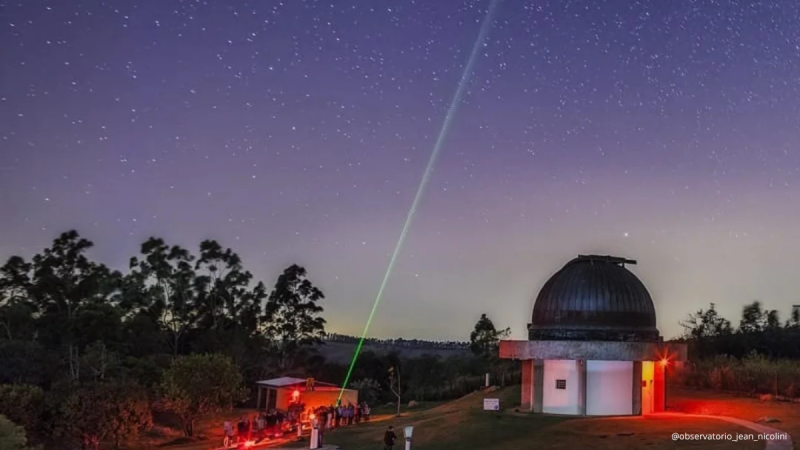 Céu noturno com estrelas e uma trilha de luz verde e roxa, em um observatório astronômico com cúpula metálica iluminada por luzes vermelhas, árvores ao fundo.