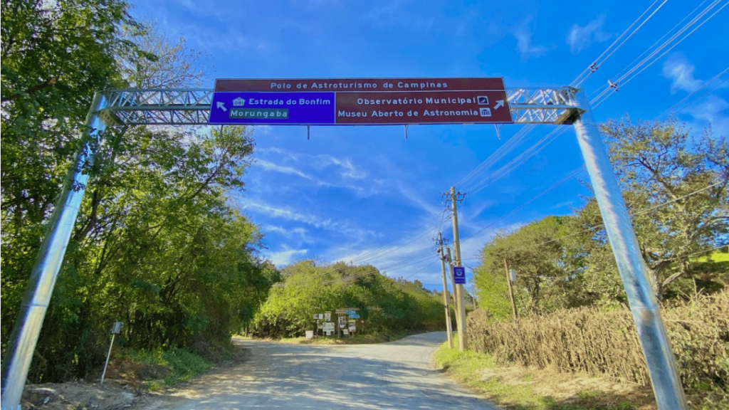 Entrada do Polo de Astroturismo de Campinas com placa de estrada, ambiente rural e vegetação ao redor, sob céu azul com árvores e postes ao longo da via.