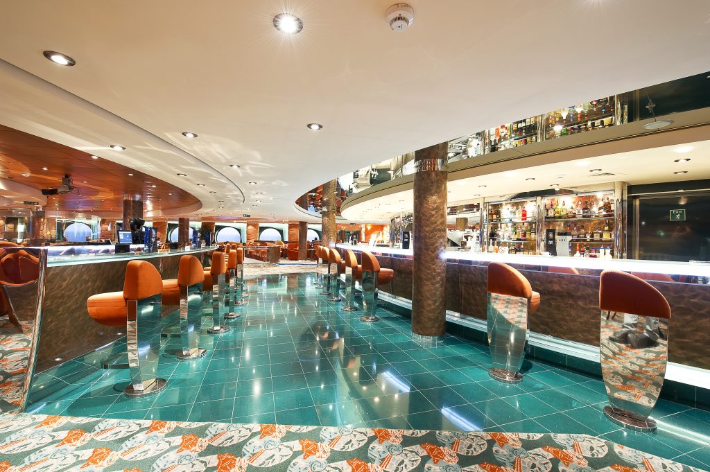 temporada de cruzeiros 2026/2027: Interior do navio de cruzeiro MSC Musica com corredor iluminado, mesas, balcões e assentos laranja na área de bar e lounge, em ambiente moderno.