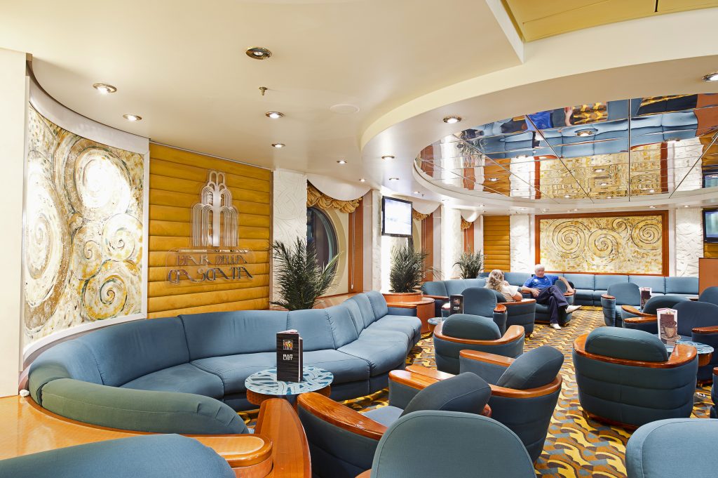 temporada de cruzeiros 2026/2027: Sala de estar em um navio de cruzeiro com assentos azuis, mesas de madeira e painéis decorativos ao fundo, em ambiente climatizado e iluminado.