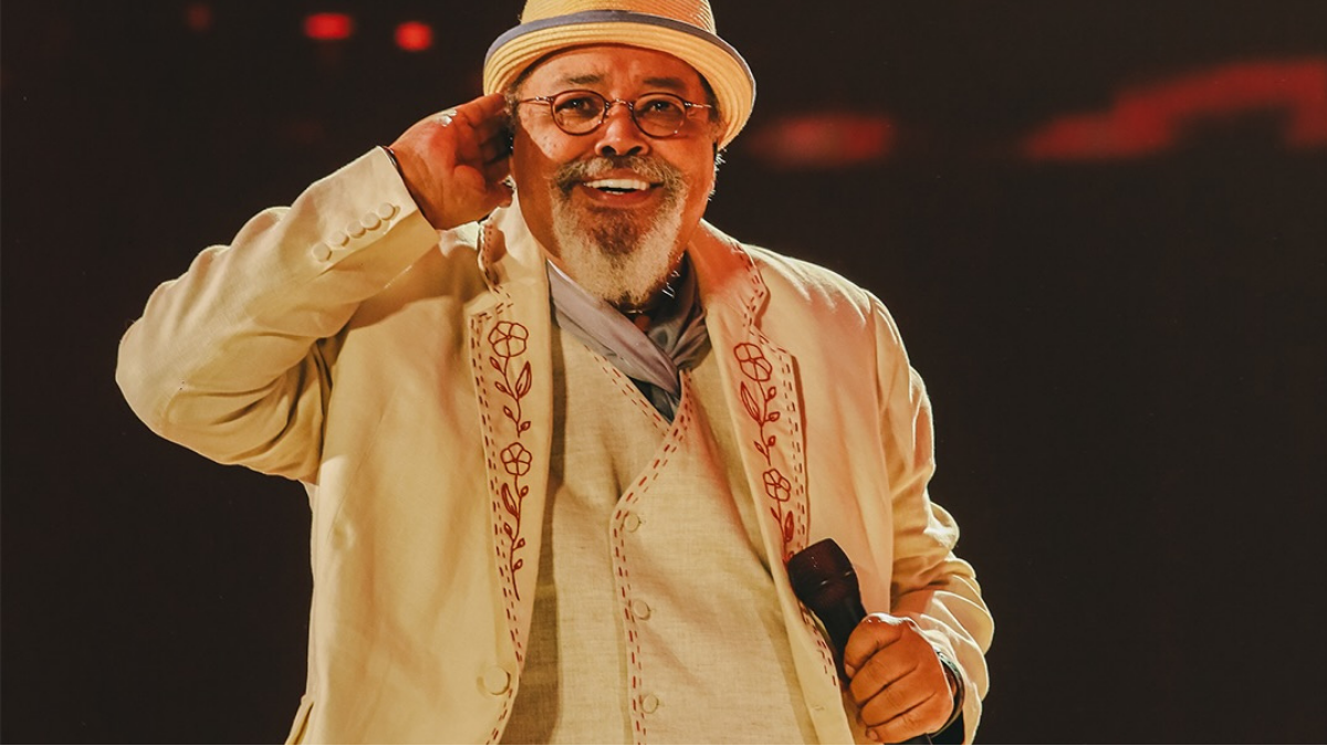 Jorge Aragão de chapéu e óculos em show, usando terno claro com detalhes bordados, segurando o microfone e fazendo gesto de braço, em palco com luzes ao fundo.