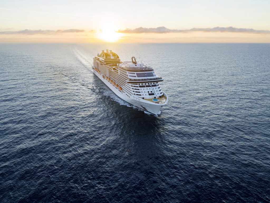 MSC Virtuosa navega pelo mar ao pôr do sol, vista aérea do navio de cruzeiro com o rastro na água, céu com nuvens e horizonte ao fundo. temporada de cruzeiros 2026/2027