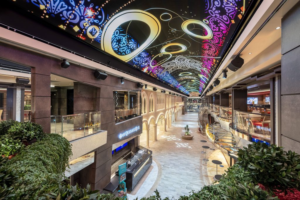 temporada de cruzeiros 2026/2027: Corredor interno do MSC Virtuosa com iluminação artística no teto e vista da Galleria Virtuosa, com lojas, áreas de descanso e vegetação decorativa