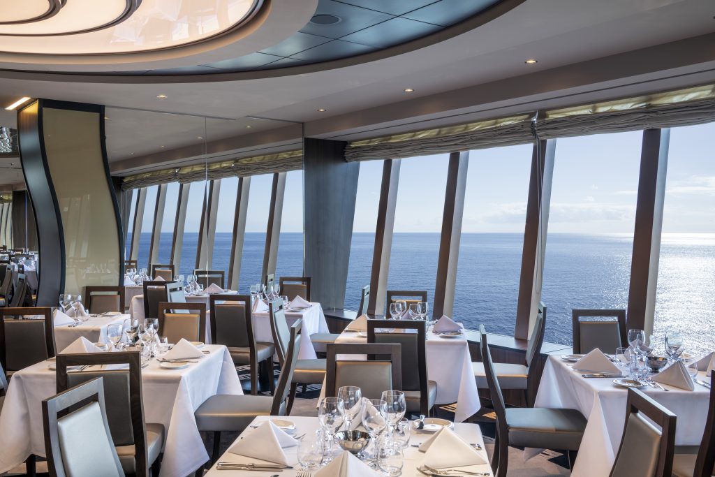 Mesa posta em restaurante com vista para o mar no MSC Virtuosa Minuetto, cenário de temporada de cruzeiros 2026/2027 com janelas panorâmicas e utensílios de jantar