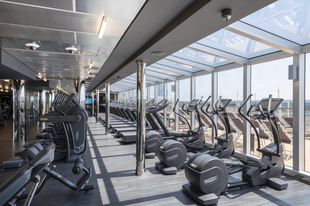 temporada de cruzeiros 2026/2027: Vista do MSC Seaview com equipamentos do MSC Gym powered by Technogym em um espaço de academia a bordo, com esteiras e aparelhos de musculação ao longo do corredor e janelas panorâmicas.