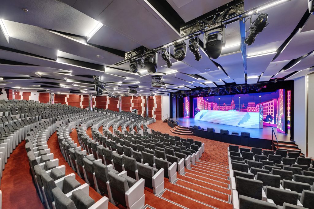 temporada de cruzeiros 2026/2027: Interior do Odeon Theatre a bordo do MSC Seaview, com fileiras de assentos e palco iluminado por luzes coloridas e cenário de evento.
