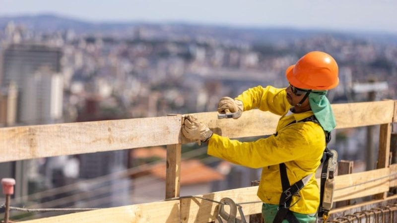 Emprego em Campinas: Trabalhador da construção civil usando capacete laranja e uniforme amarelo, realizando instalação em andaime alto com a cidade ao fundo.