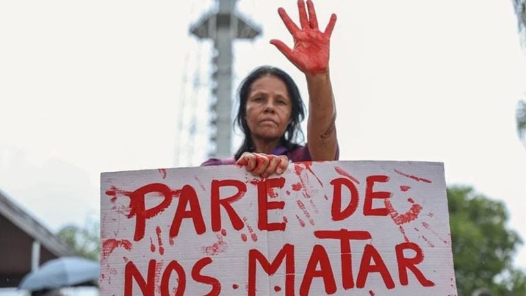 Lei Maria da Penha 20 anos: Manifestação contra a violência doméstica: mulher ergue a mão e segura um cartaz escrito “PARE DE NOS MATAR”, com cenário ao ar livre em homenagem aos 20 anos da Lei Maria da Penha