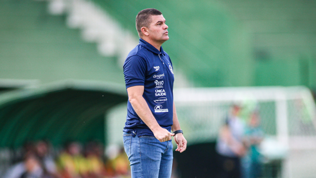 Umberto Louzer, técnico do Guarani, em pé no campo com expressão concentrada durante partida de futebol, usando uniforme azul e observando o jogo ao fundo.