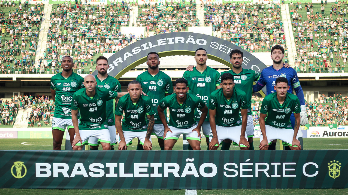 Elenco do Guarani em foto de apresentação no estádio, com uniforme verde e técnico e jogadores atrás de faixa do Brasileirão Série C, após eliminação
