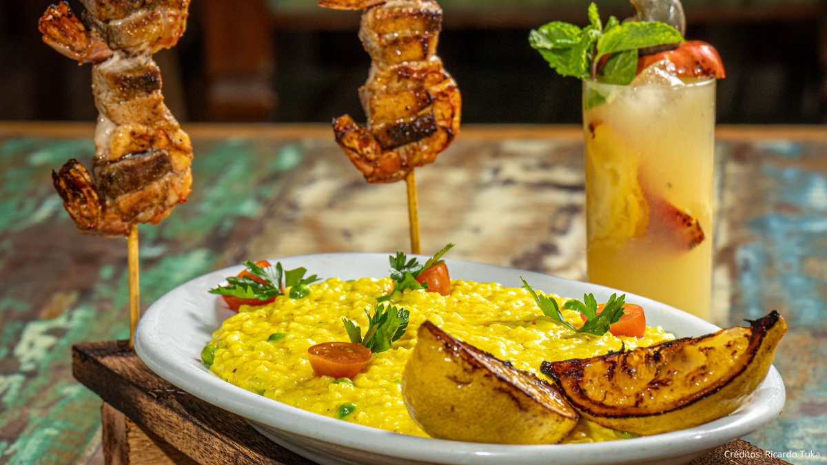 Prato com arroz amarelo cremoso, fatias de abóbora grelhada e espetinhos de carne na bancada, ao lado de um drink em copo alto com gelo e fatia de fruta, em ambiente de restaurante.