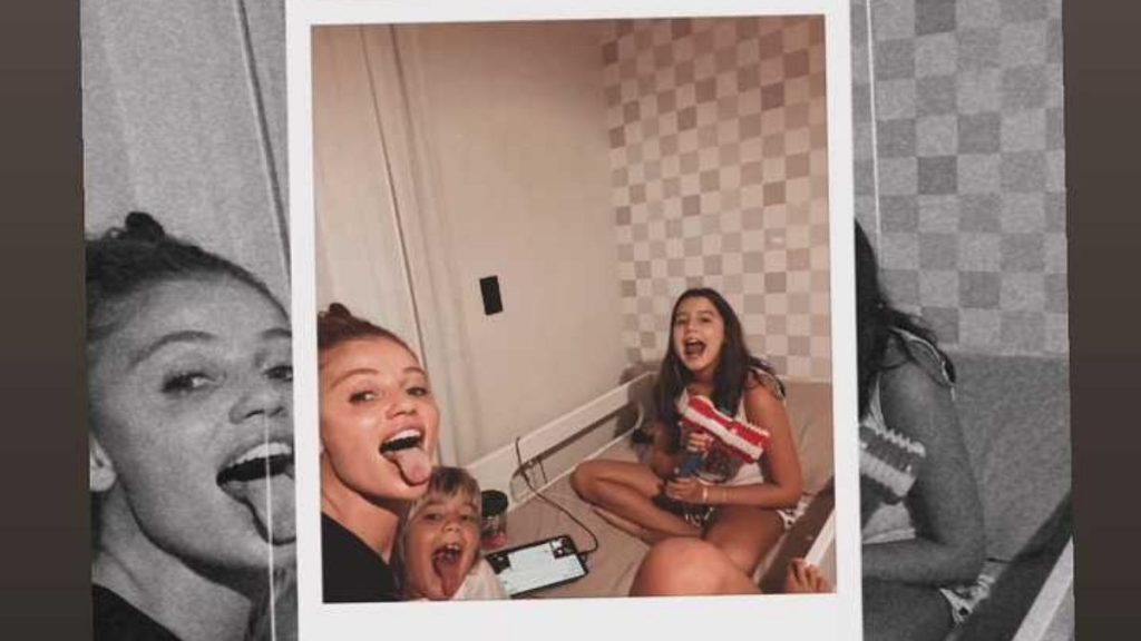 Foto em estilo polaroid mostrando uma mulher cantando com um microfone e uma menina ao lado, enquanto outra criança segura um controle de brinquedo em um ambiente interno.