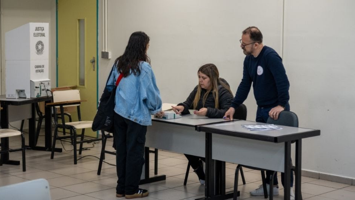 Pessoa em sala de atendimento com mesa de votação, agentes orientando eleitora e urna eletrônica ao fundo, em ambiente de justiça eleitoral.
