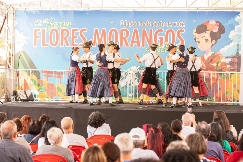 Divulgação da Festa de Flores e Morangos de Atibaia, com grupo folclórico dançando no palco durante evento ao ar livre, em cenário decorado