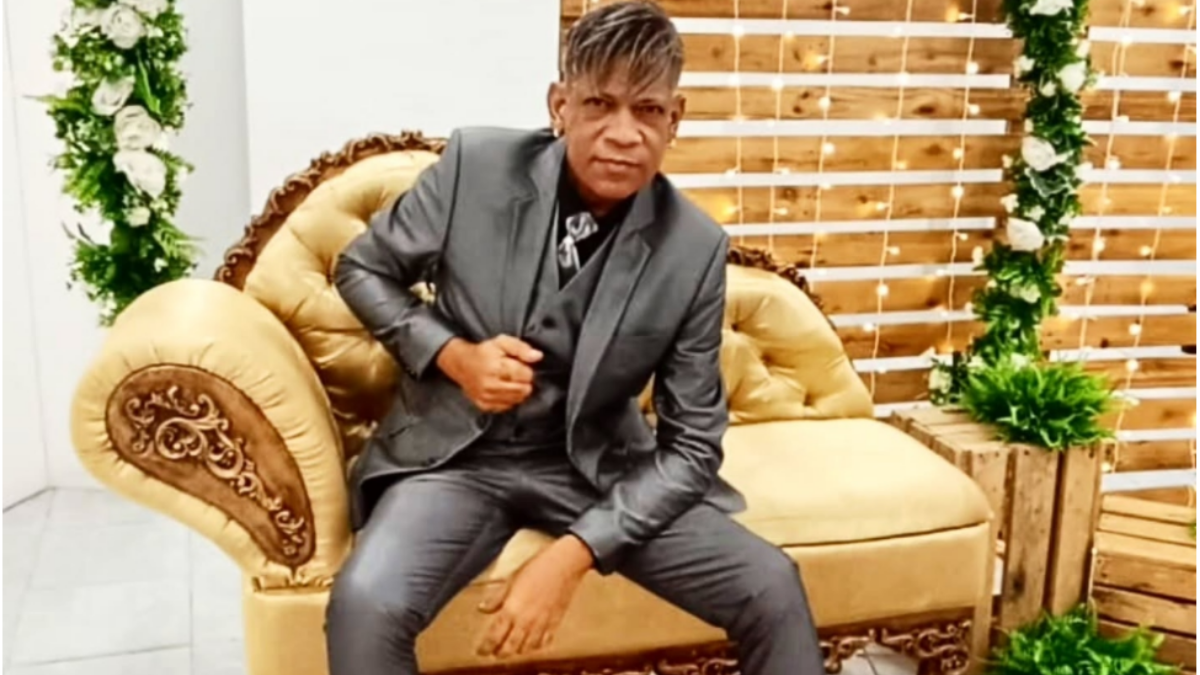 Homem adulto em um sofá bege com detalhes dourados, usando terno escuro, sentado em ambiente decorado com folhagens e luzes decorativas atrás.