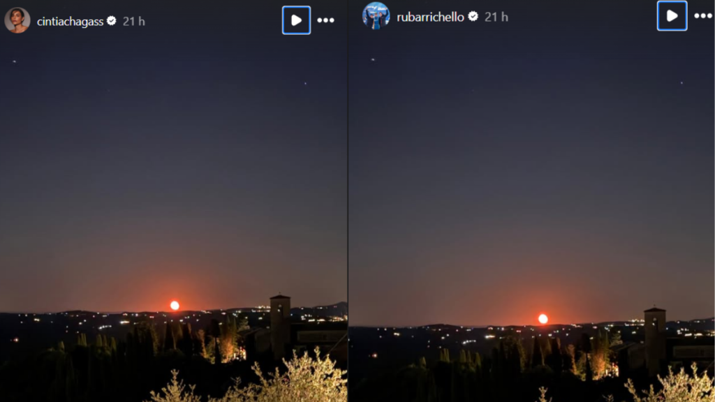Stories Cíntia Chagas e Rubens Barrichello, foto céu noturno com gradiente em tons de azul e laranja ao amanhecer, luzes da cidade ao fundo e árvores iluminadas em primeiro plano, com poste e prédio ao lado.