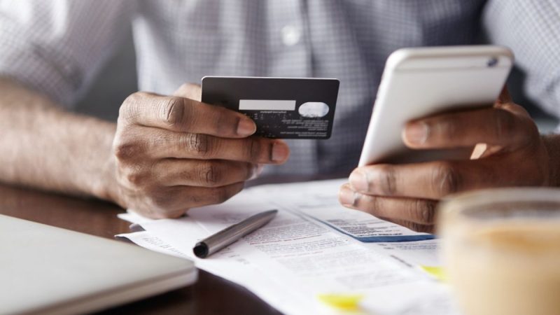 Pix tem novas regras contra fraudes: pessoa segura cartão bancário e celular durante transação, com documentos e caneta sobre a mesa, sugerindo segurança no pagamento.