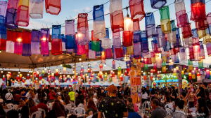 Decoração de festa com lanternas coloridas penduradas e luzes ao fundo, com pessoas reunidas em um ambiente de evento ao ar livre.