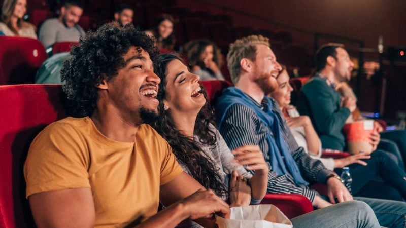 Casal e amigos sentados em poltronas de cinema, sorrindo durante a sessão; destaque para o clima de diversão em de cinema escura em alusão a Semana do Cinema.