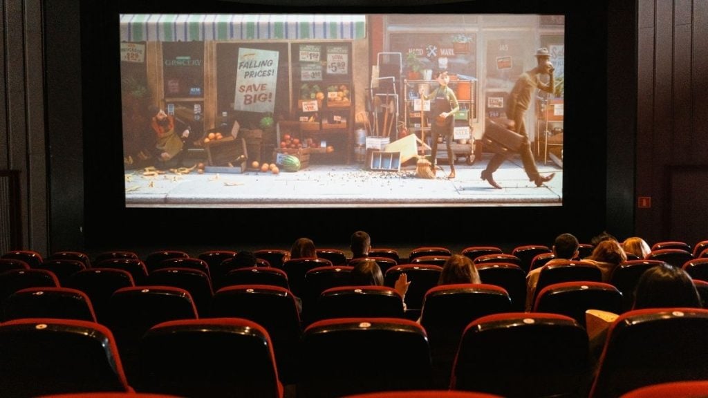 Sessão de cinema em uma sala escura com fileiras de cadeiras vermelhas e uma tela exibindo um filme com cenário urbano e personagens em frente a uma banca.