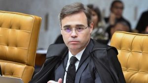 Moraes Vorcaro: André Mendonça em sessão no tribunal, ao centro, com toga e faixa, sentado em cadeira amarela, imagem desfocada ao fundo; tema envolve sigilo de conversas entre Moraes e Daniel Vorcaro.