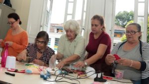 Cursos de artesanato em Santos: Aula em grupo com participantes sentadas ao redor de uma mesa, criando artesanato com materiais e linhas, em divulgação de Santos abre inscrições para cursos de artesanato, maquiagem e padaria.