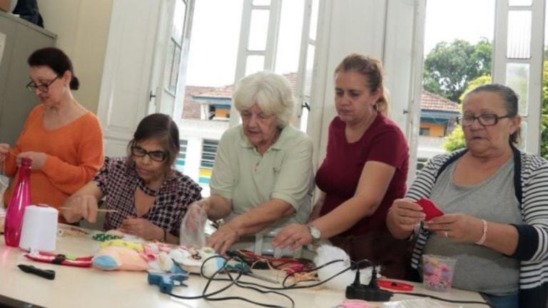 Cursos de artesanato em Santos: Aula em grupo com participantes sentadas ao redor de uma mesa, criando artesanato com materiais e linhas, em divulgação de Santos abre inscrições para cursos de artesanato, maquiagem e padaria.