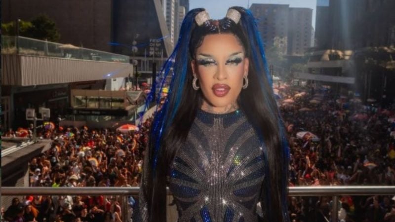 Gloria Groove aparece em um evento ao ar livre, com cabelo longo e azul e maquiagem artística, vestindo um look brilhante escuro, em meio a uma multidão ao fundo.