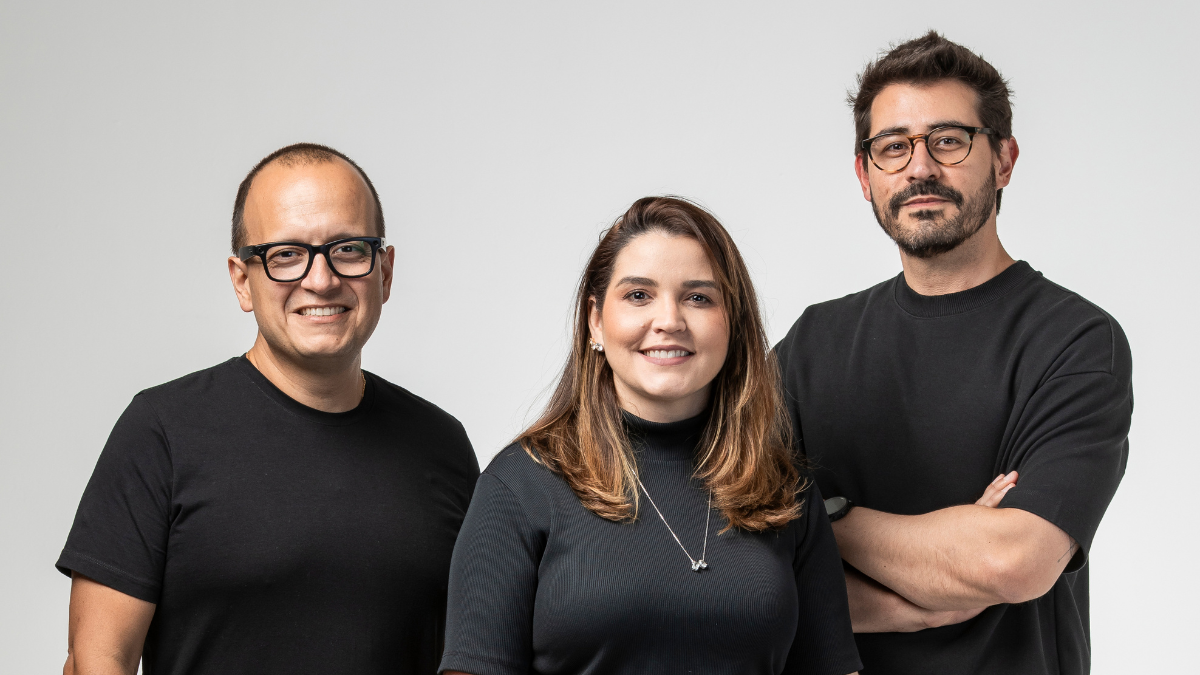 Equipe de três pessoas em estúdio com fundo claro: um homem de óculos à esquerda, uma mulher sorridente no centro e outro homem de óculos à direita, em foto corporativa