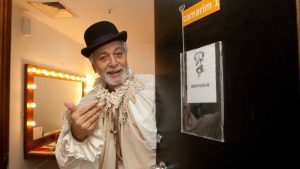 Gracindo Jr. em foto de bastidores, com chapéu e barba, em um corredor de produção ao lado de uma porta com identificação “Camarim 1”.