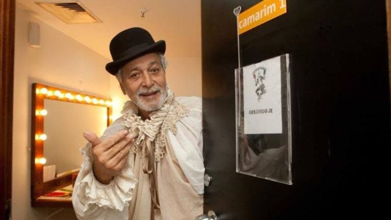 Gracindo Jr. em foto de bastidores, com chapéu e barba, em um corredor de produção ao lado de uma porta com identificação “Camarim 1”.