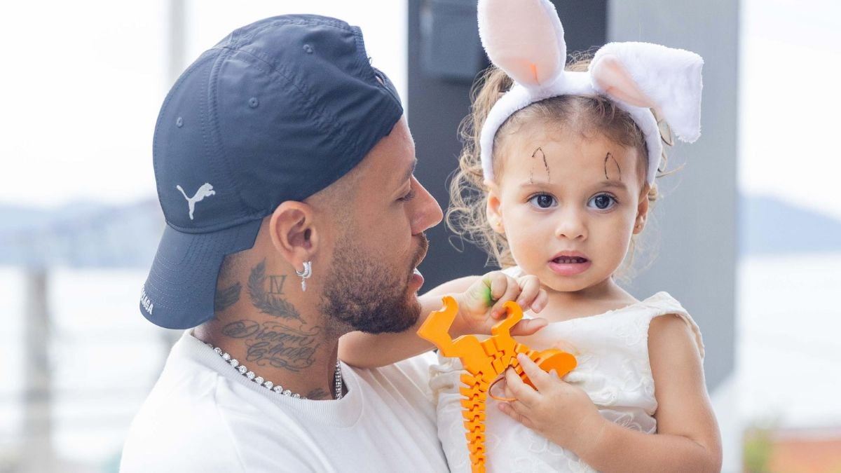 Neymar e filha helena: Neymar aparece ao lado de sua filha Helena, uma criança usando touca de coelho, repetindo a mesma careta enquanto o pai a observa sorrindo em um momento de carinho.