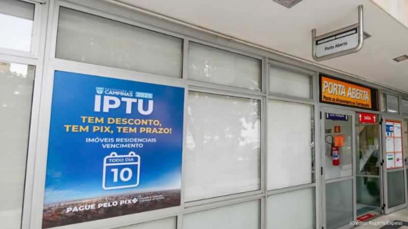 Entrada de prédio da Prefeitura de Campinas com placas de anúncio de promoções do IPTU, incluindo destaque “PORTA ABERTA”, portas de acesso e janelas de vidro ao lado.