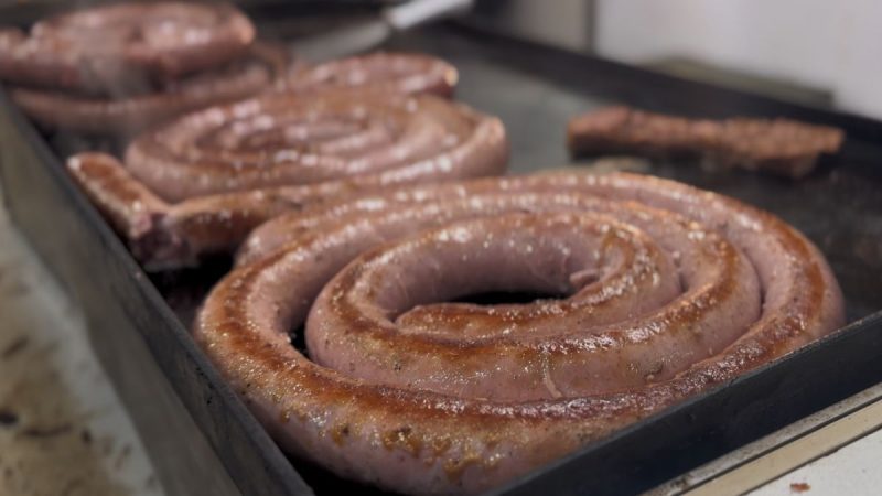 Linguiças assadas em uma grelha, com o Festival da Linguiça de Bragança Paulista ao fundo sugerido pela cena de preparo de salsichas na churrasqueira.