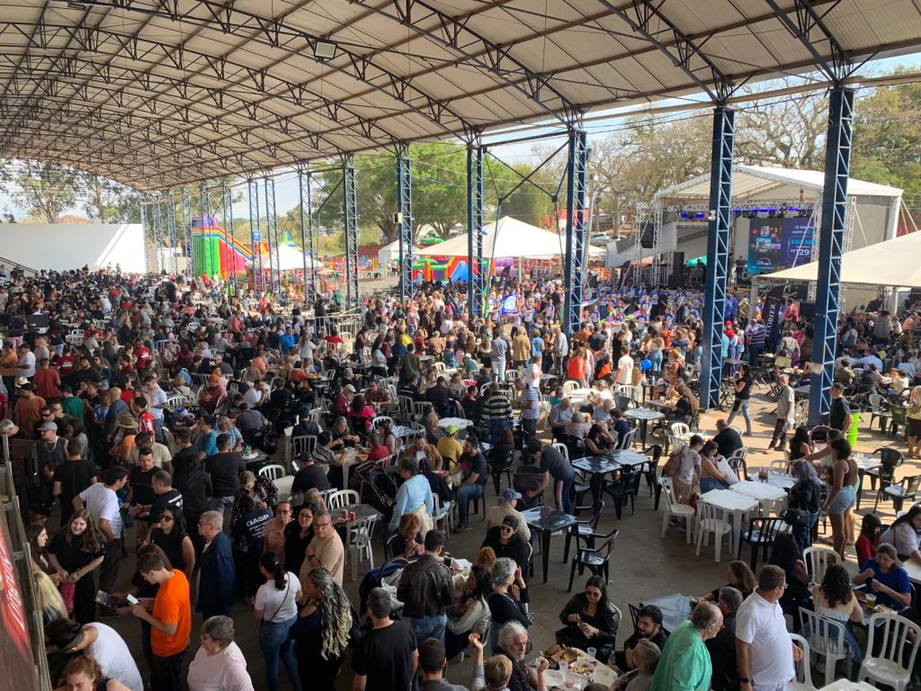 Multidão reunida em um grande evento ao ar livre sob tendas e palco, com barracas e mesas ao fundo, em clima de Festival da Linguiça de Bragança Paulista.