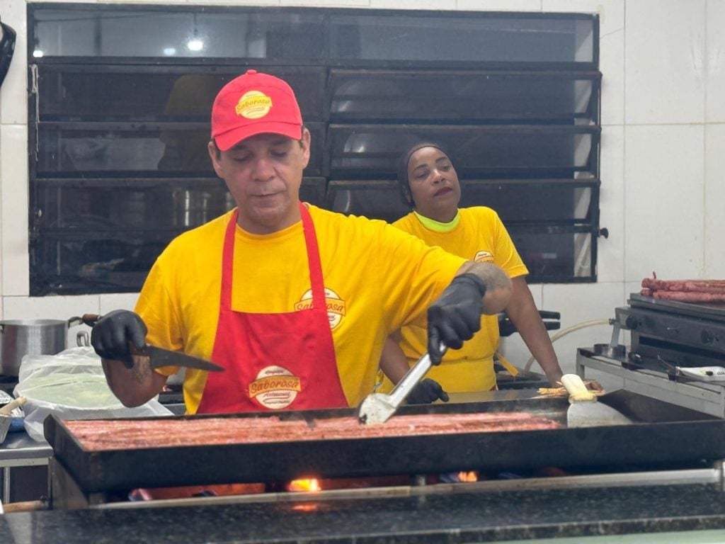 Festival da Linguiça de Bragança Paulista: Cozinheiro em churrasqueira industrial prepara linguiça no grelhador, usando touca e avental vermelho, enquanto outra pessoa ao fundo acompanha a produção em evento.