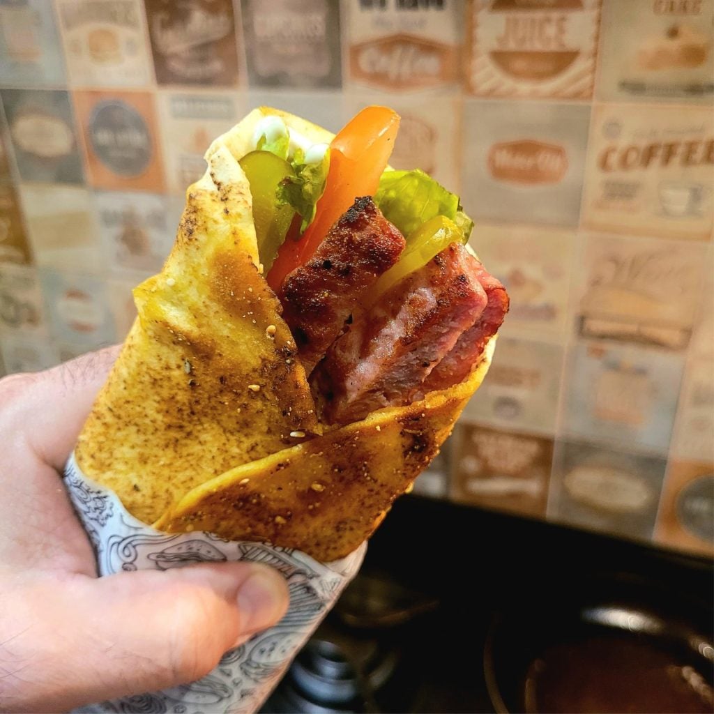 Festival da Linguiça de Bragança Paulista: sanduíche de linguiça na mão, com batata frita crocante e molhos, ao fundo um painel de publicidade desfocado.
