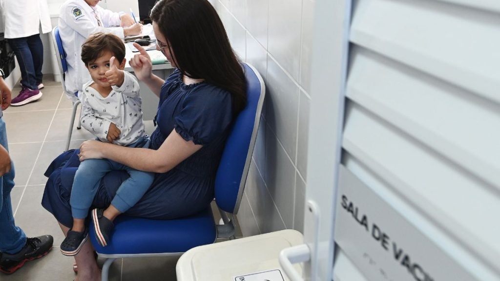 Profissional de saúde aplica vacina em criança sentada em cadeira durante campanha de vacinação contra gripe em campinas, em clínica com ambiente de atendimento